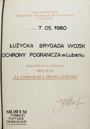 1. Tytuł: postanowienie o nadaniu medalu „Za zasługi dla miasta Lubania”
2. Treść: postanowienie Prezydium Miejskiej Rady Narodowej w Lubaniu z dnia 07.05.1980 r. o wyróżnieniu medalem „Za zasługi dla miasta Lubania” Łużyckiej Brygady WOP 
3. Hasło przedmiotowe: Wojska Ochrony Pogranicza, przedmioty pamiątkowe
4. Wytwórca: Prezydium Miejskiej Rady Narodowej w Lubaniu
5. Czas powstania: 07.05.1980 r.
6. Miejsce sporządzenia: Polska, Lubań
7. Materiał i technika wykonania: papier, druk wypełniony pismem odręcznym
8. Wymiary: 30 x 21 cm
9. Stan zachowania: dobry
10. Miejsce przechowywania/ właściciel: depozyt Sali Tradycji Ośrodka Szkoleń  Specjalistycznych SG w Lubaniu